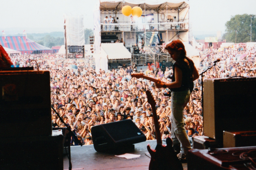 A teenage boy on stage holding a guitar in front of a large festival crowd