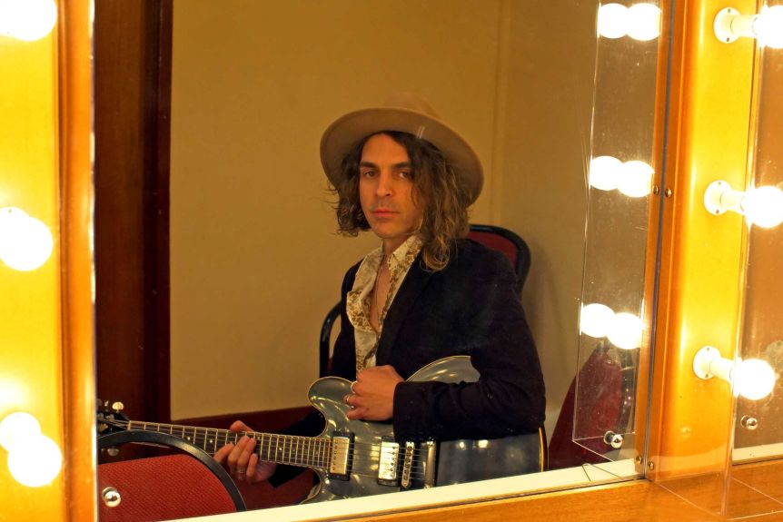 A portrait of a man holding a guitar and wearing a wide brim hat reflected in a mirror that's lined with light bulbs